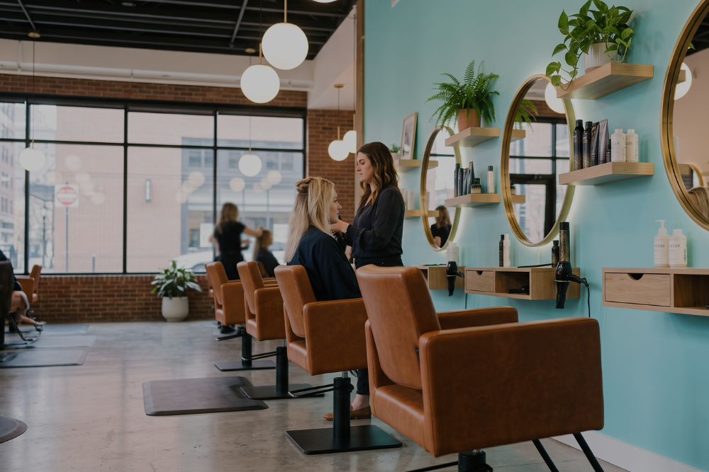 A modern salon interior with several brown leather styling chairs, round gold-framed mirrors, wall shelves, and hanging white globe lights against a turquoise wall. Two women are standing near the salon station, with one seated client visible in the background near the window.